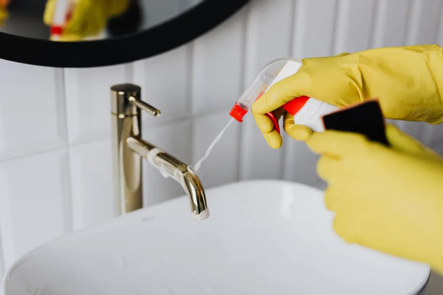 Cypress - From above anonymous housekeeper in yellow rubber gloves spraying cleaner to chrome water faucet in bathroom